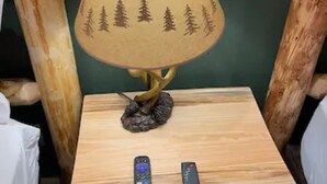 A wooden bedside table with a lamp featuring a pine tree design, two remote controls, and a green wall in the background. - Cedar Mountain - Suite B (Grants Pass)