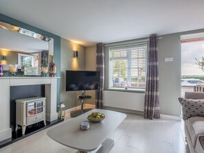 A modern living room with a TV, a fireplace, a dining table with a bowl of fruit, and a large window with curtains.
