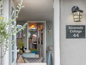 Une entrée accueillante, avec une pancarte indiquant « Riverside Cottage 44 », un coin repas avec une table dressée pour deux personnes, et un escalier menant à l’étage supérieur.