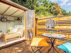 Une terrasse extérieure avec un canapé en osier, une table ronde sur laquelle reposent deux verres de vin rouge, et une clôture en bois ornée d’un motif représentant un arbre.