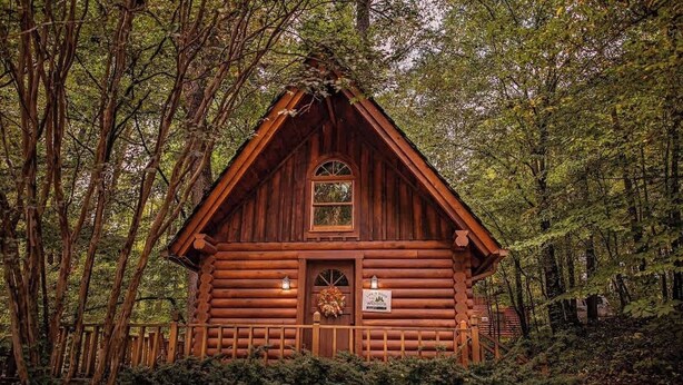 A log cabin with a pitched roof, a central door, and two lit lanterns on either side. - Ivy quaint tiny cabin creekside - Riverside Resort (Benton)
