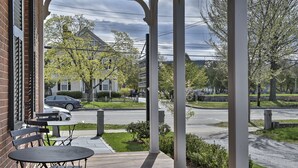A covered outdoor seating area with tables and chairs, overlooking a residential street with houses and a road. - The Burrell House (Keene)