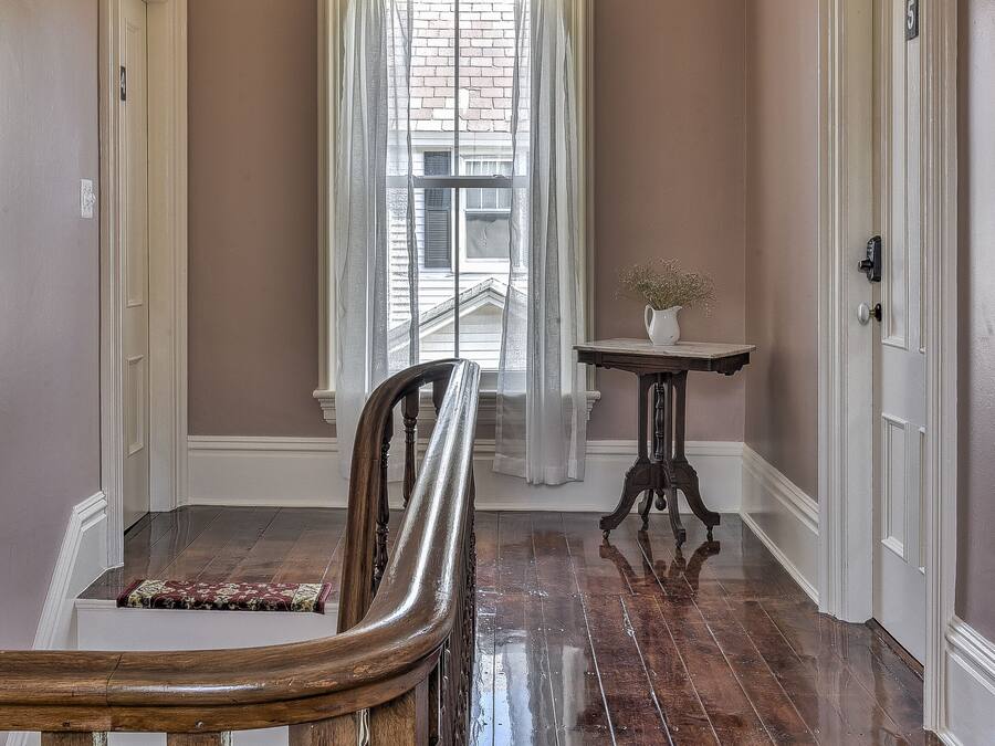 Un pasillo con escaleras de madera, una mesa con un florero y una ventana con cortinas traslúcidas.