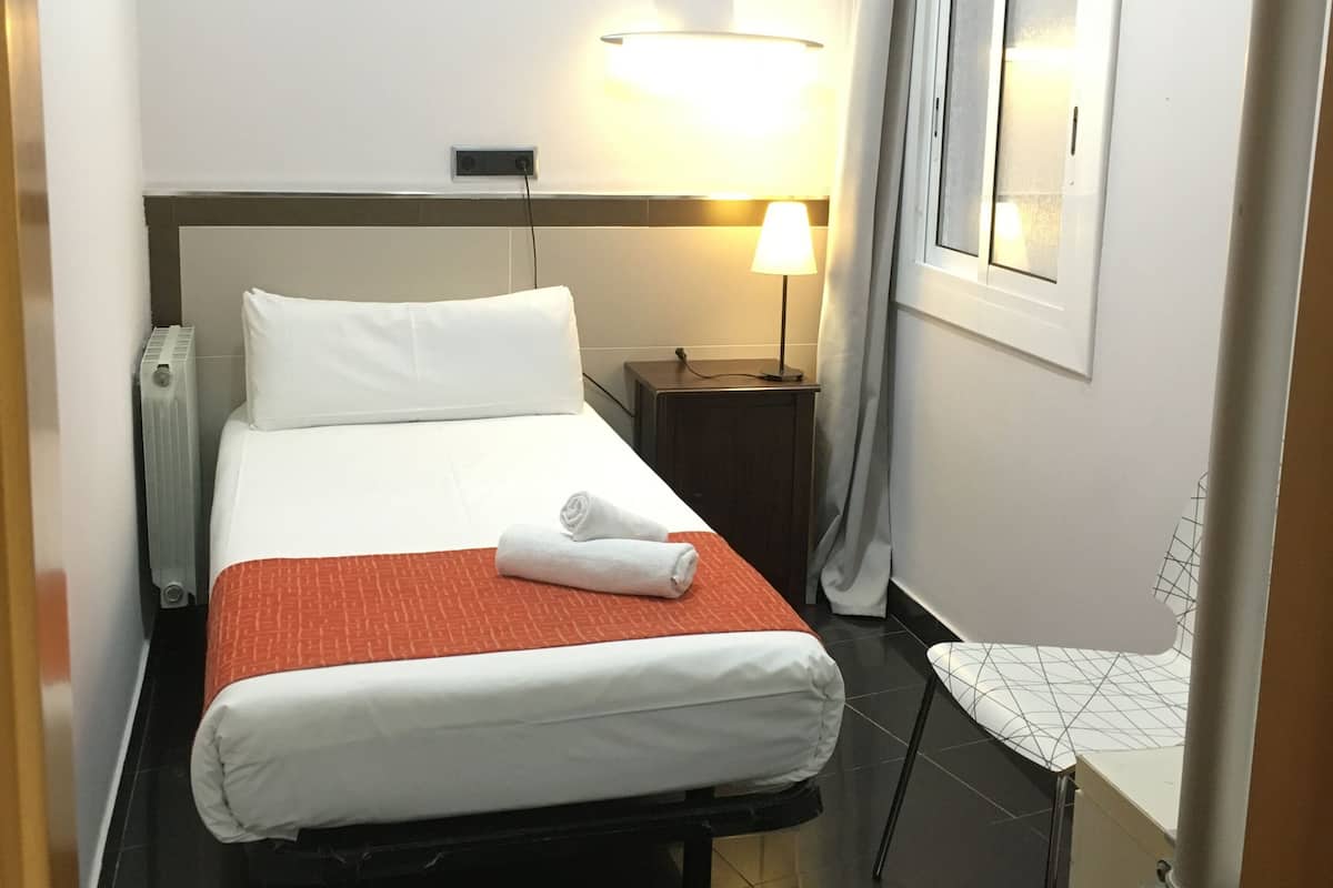 A hotel room with a bed, a bedside table, a lamp, a radiator, and a window with curtains.