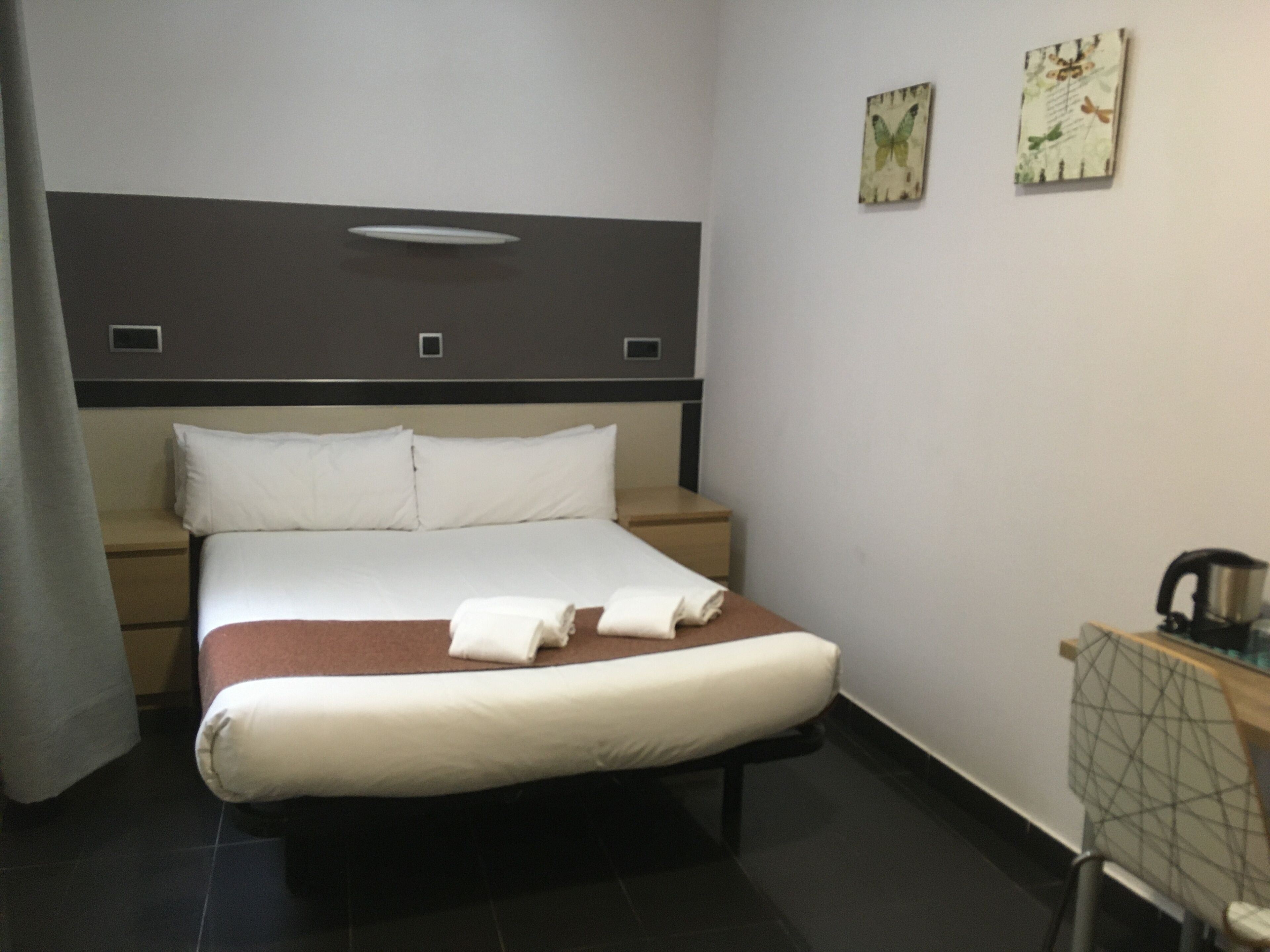 A hotel room with a bed, bedside tables, a nightstand, a desk with a chair, and a wall-mounted shelf.