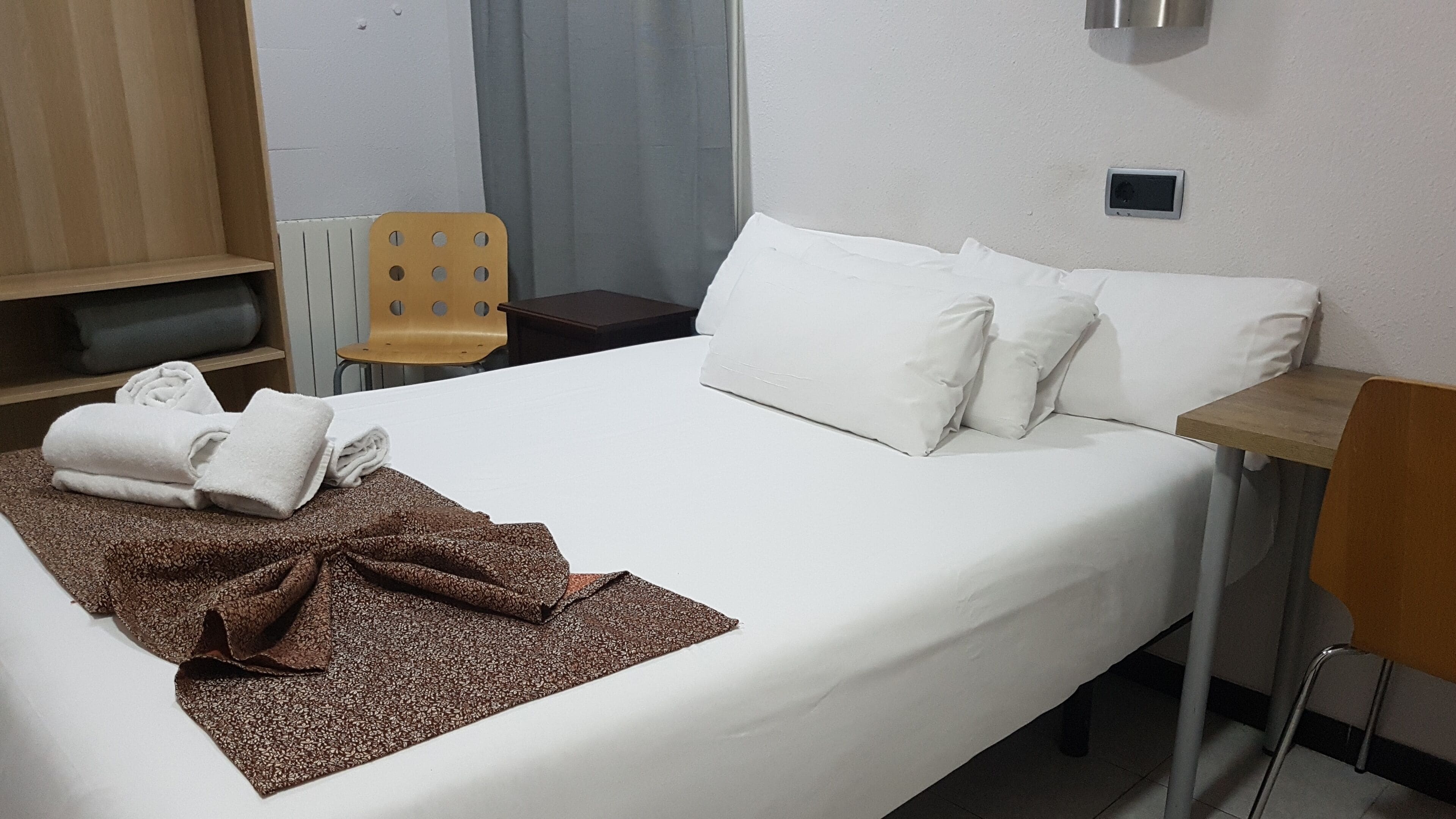 Una cama bien hecha con sábanas y almohadones blancos, una manta marrón estampada y toallas enrolladas sobre un estante de madera.