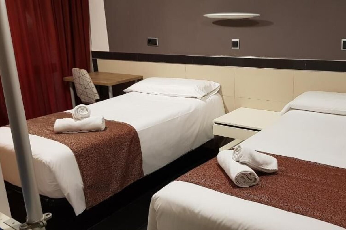 A hotel room with two single beds, each with a brown headboard, white linens, and a rolled towel on the bed. There is a small bedside table with a lamp and a wall-mounted light fixture above the beds.