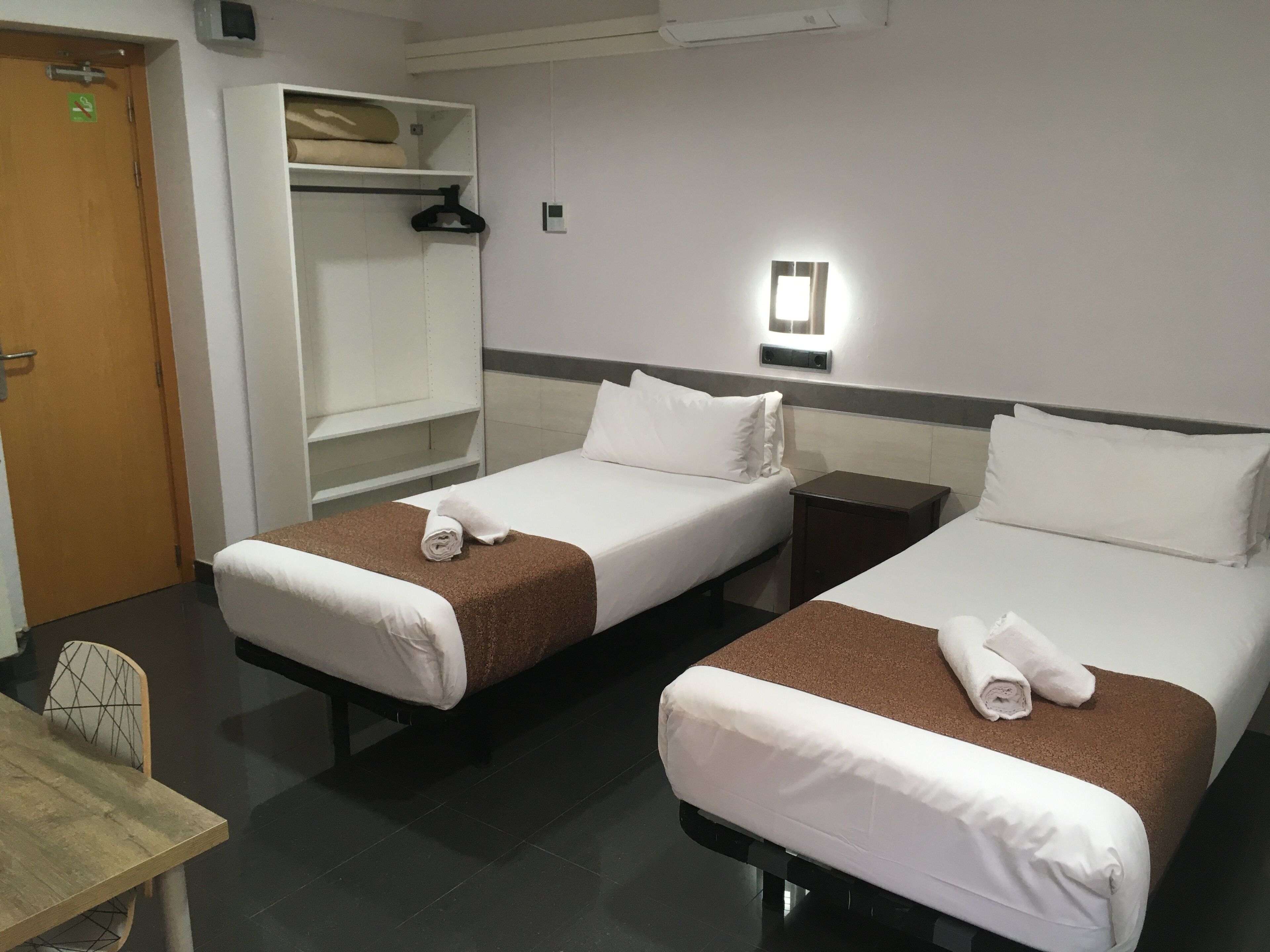 A hotel room with two single beds, a wooden table, a wardrobe, and a wall-mounted light fixture.