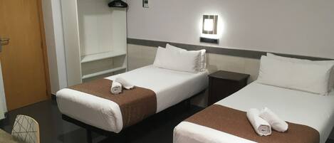 A hotel room with two single beds, a wooden table, a wardrobe, and a wall-mounted light fixture.