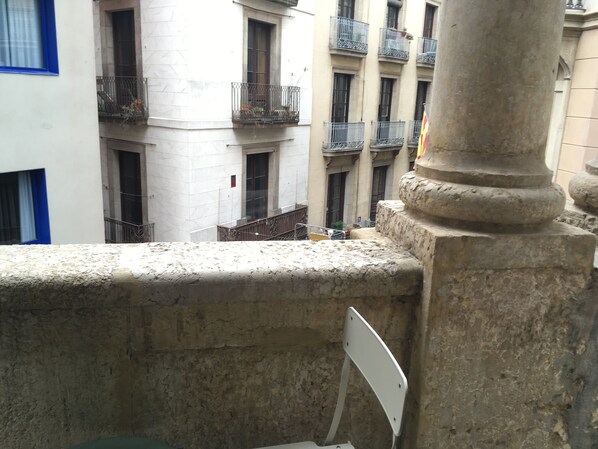 Una barandilla de piedra con una silla de metal, un edificio blanco con ventanas de marco azul, y un edificio con varios balcones.