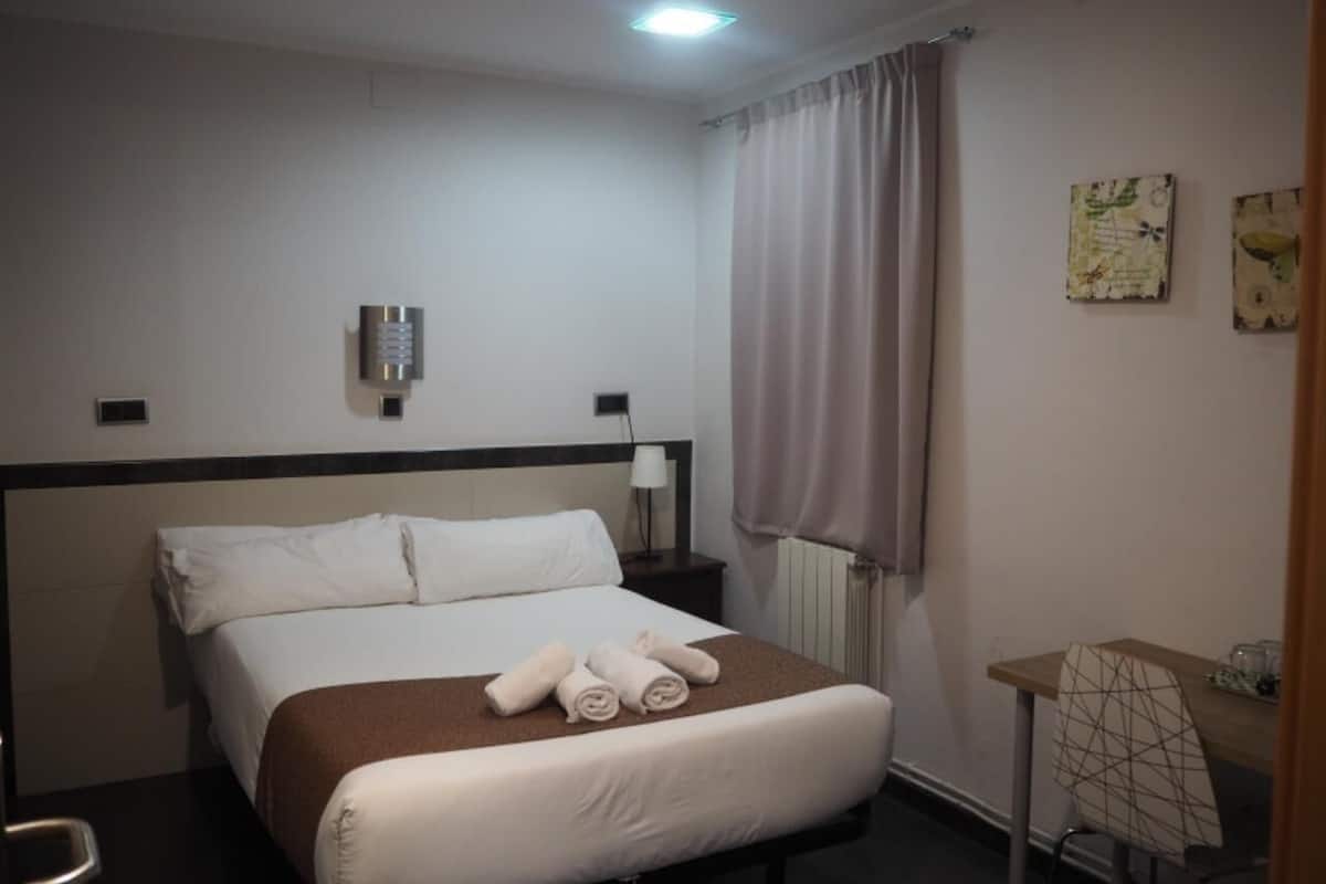 A hotel room with a bed, a desk, a chair, a lamp, and a window with curtains.