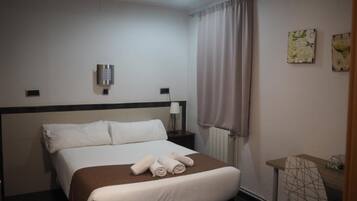 A hotel room with a bed, a desk, a chair, a lamp, and a window with curtains.