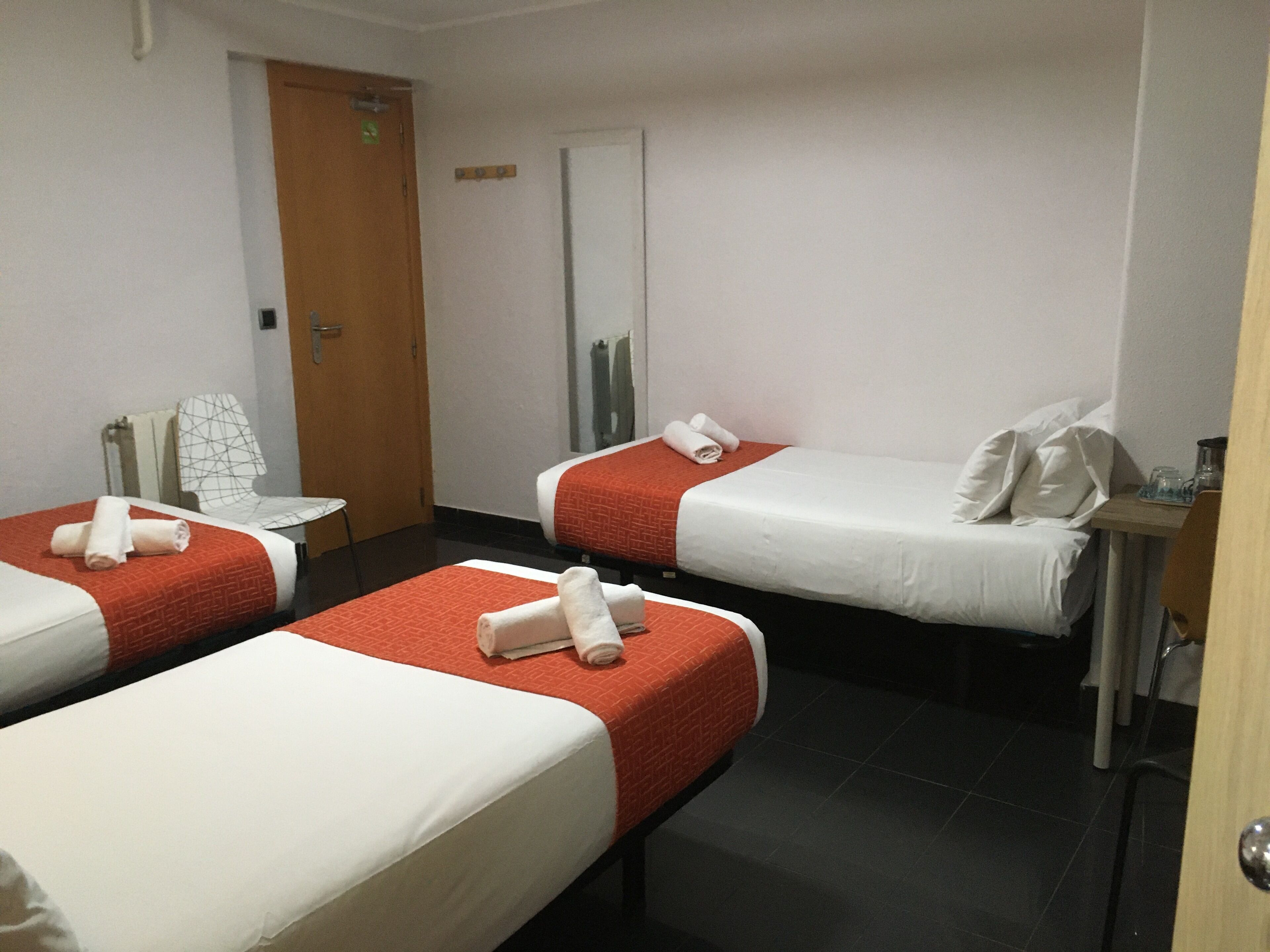 Ein Hotelzimmer mit drei Einzelbetten, jeweils mit einer roten und weißen Bettdecke und ordentlich gefalteten Handtüchern. Man sieht eine Holztür, einen kleinen Tisch mit einem Stuhl und einen Spiegel an der Wand.