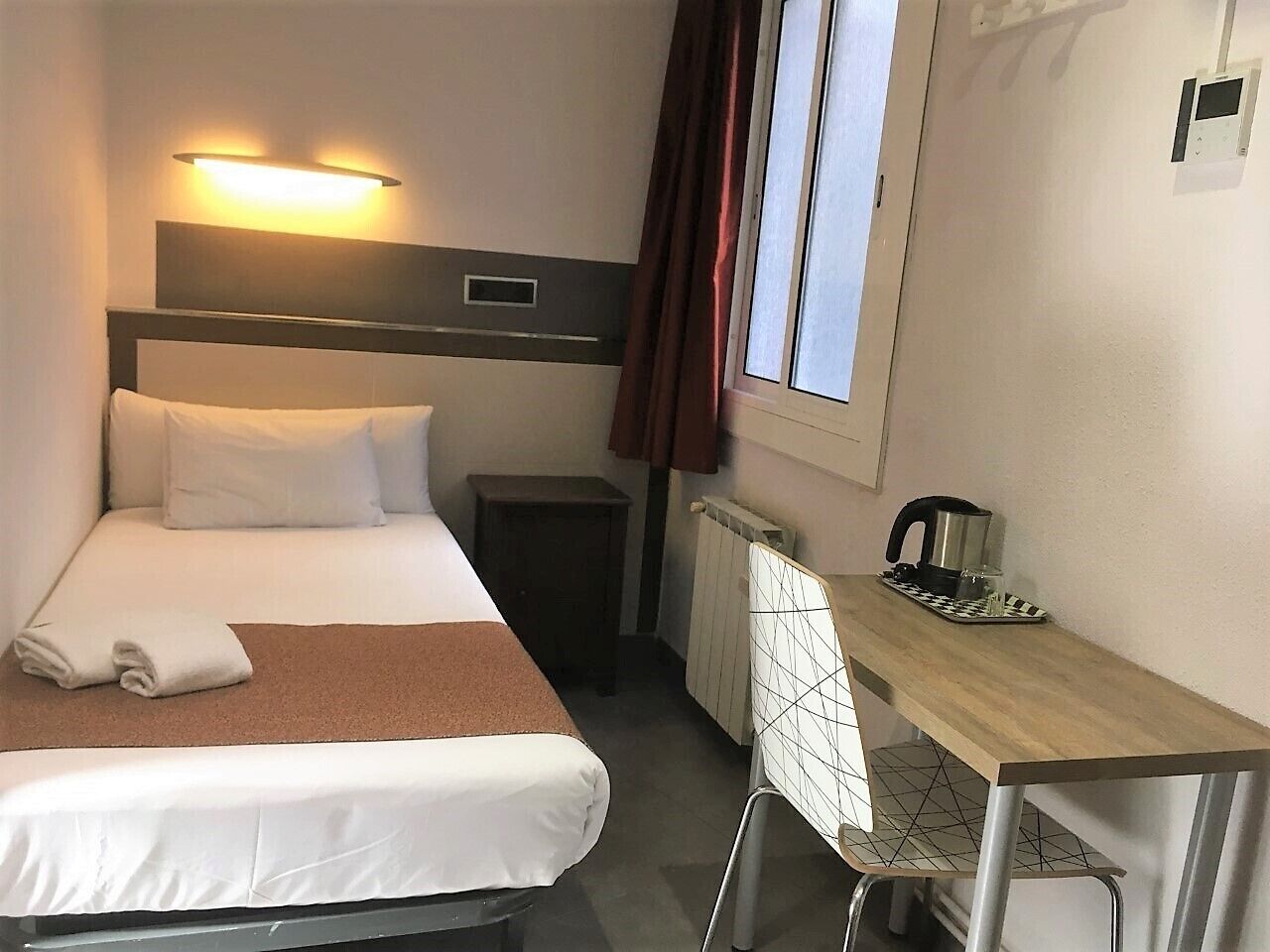 A hotel room with a bed, a desk with a kettle, a radiator, and a window with curtains.