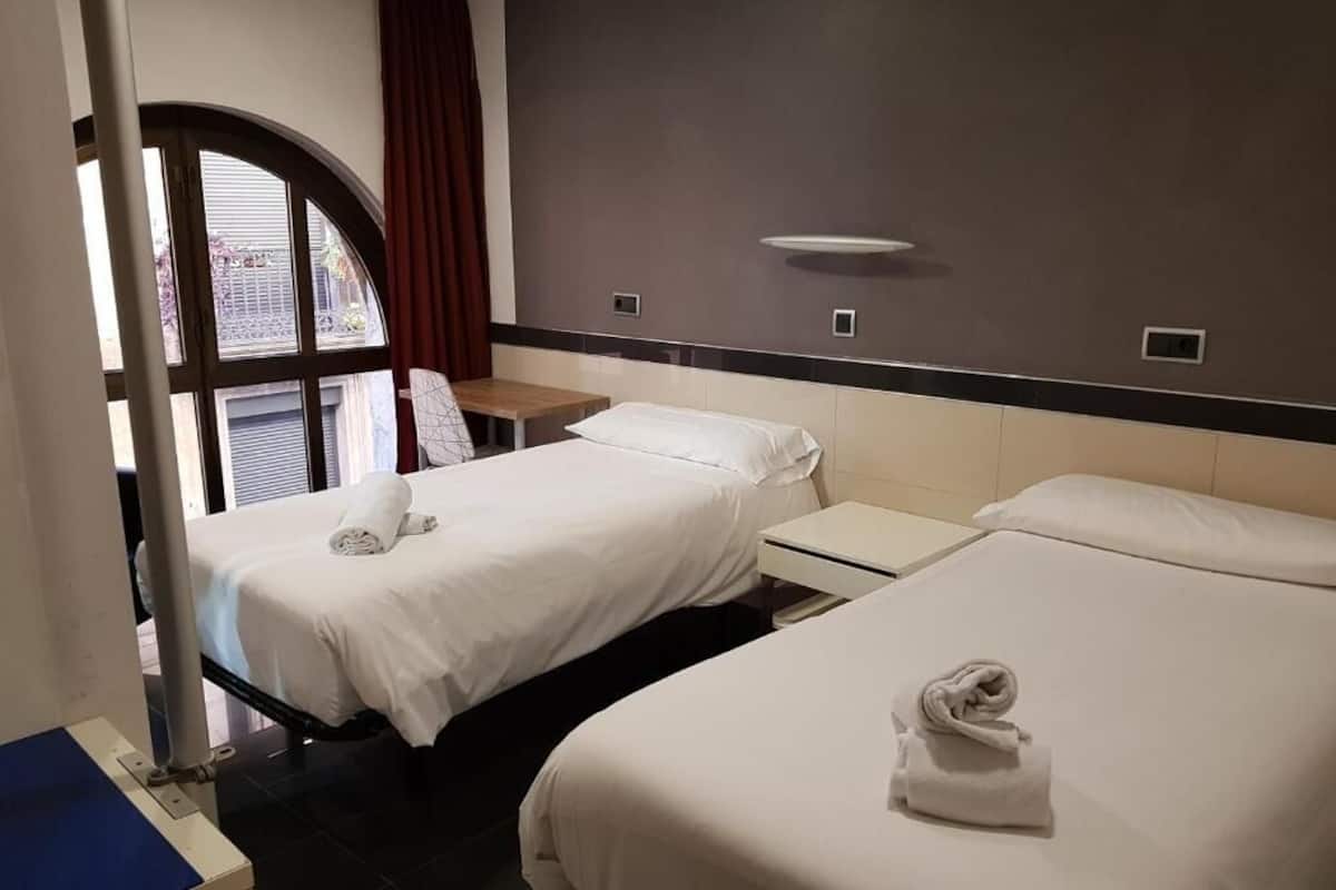 A hotel room with two beds, a small table, and a window with curtains.