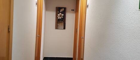 A narrow hallway with black tiled flooring, white walls, and wooden doors on both sides. There is a number '108' on the wall and a framed picture of flowers.