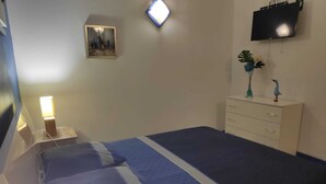 Un dormitorio con cama, mesita de noche, lámpara, televisor y cómoda con objetos decorativos.