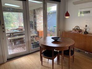A wooden dining table with chairs, a glass door leading to an outdoor patio, and a wooden cabinet with a lamp. - Somers Shack - walk to beach through Koala Reserve (Somers)