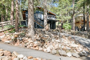 Eine moderne Holzhütte mit einer Holzdiele und Treppen, die zu einer zweiten Ebene führen, umgeben von felsigem Gelände und dichtem Baumbestand.