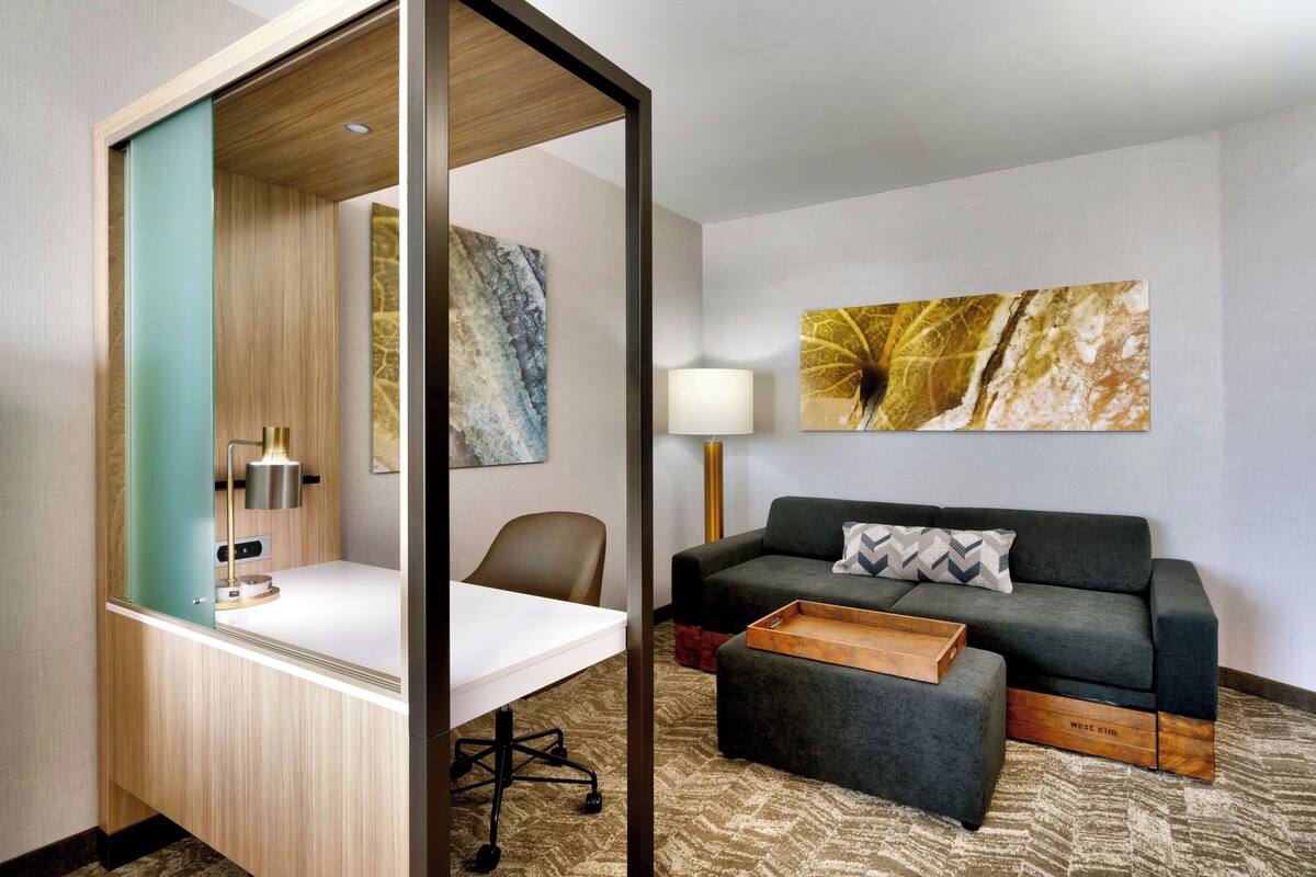 A modern hotel room with a desk, chair, sofa, and ottoman.