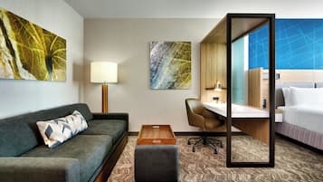 A hotel room with a grey sofa, a wooden side table, a desk with a chair, a bed, and two artworks on the walls.