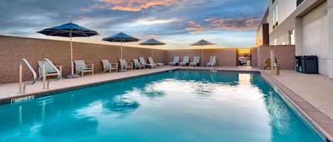 Una piscina con sillas de descanso y sombrillas, rodeada por un muro de ladrillo y un edificio.