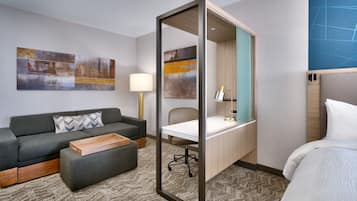 A hotel room with a grey sofa, a wooden side table, a desk with a chair, a bed, and abstract wall art.