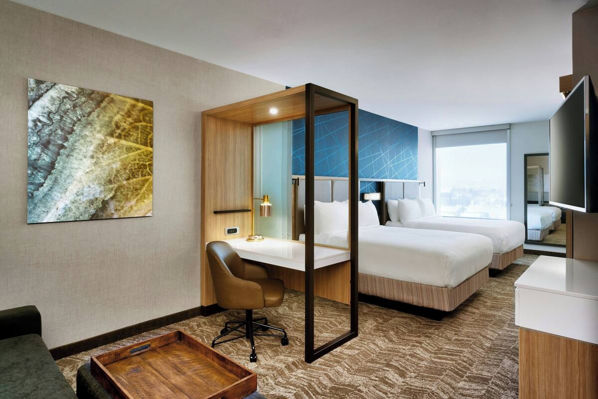 A hotel room with a desk, two beds, a large window, and a city view.
