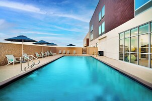 Una piscina con sillas de descanso y sombrillas, rodeada por un edificio con ventanales amplios.