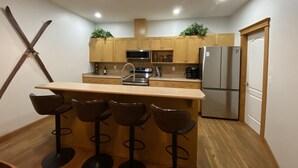 A modern kitchen with wooden cabinets, a stainless steel refrigerator, and a bar area with stools. - Lg Townhouse (Bigwhite)
