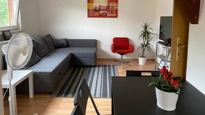 Una sala moderna con un sofá seccional gris, una silla roja, una mesa de comedor con sillas, un ventilador, una planta en maceta y un cuadro en la pared.