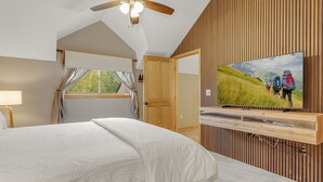 Quarto com cama, ventoinha no teto, janela com cortinas, porta de madeira e televisão fixada em parede de madeira.