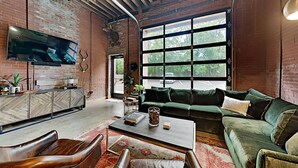 A spacious living room with a large window, a sofa, a coffee table, and a TV mounted on the wall. - One-Of-A-Kind! Roll Up Garage Door, Pool, Speakeasy (Nashville)