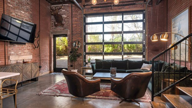 A spacious room with exposed brick walls, a large window with a view of greenery, a black leather sofa, a coffee table, and a TV mounted on the wall. - One-Of-A-Kind! Roll Up Garage Door, Pool, Speakeasy (Nashville)