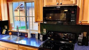 A kitchen with wooden cabinets, a black microwave, a stove, and a window with a view of a wooden cabin. - Cedar Mountain - Suite E (Grants Pass)