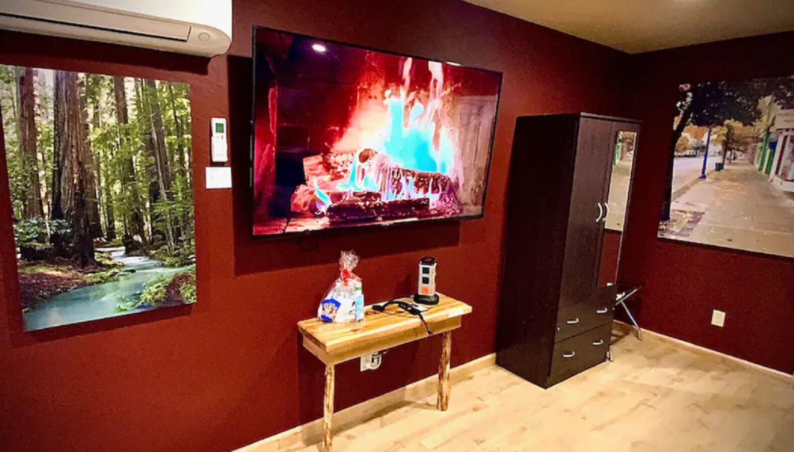 Ein Zimmer mit an der Wand befestigtem Fernseher, der eine Feuerszene zeigt, einem kleinen Tisch mit einer Tasche und einem Gerät sowie einem Waldposter.