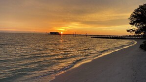 Une plage avec une jetée en bois qui s’avance dans l’eau, un coucher de soleil en arrière-plan et un arbre sur la droite.
