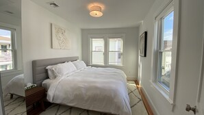 Un dormitorio con cama, mesita de noche, una obra de arte en la pared y una ventana con persianas.