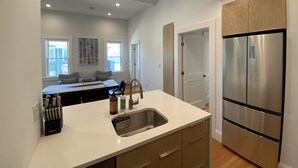 Una cocina moderna con un refrigerador de acero inoxidable, una encimera blanca y un lavamanos. Se ve un área de comedor al fondo con una mesa y sillas.