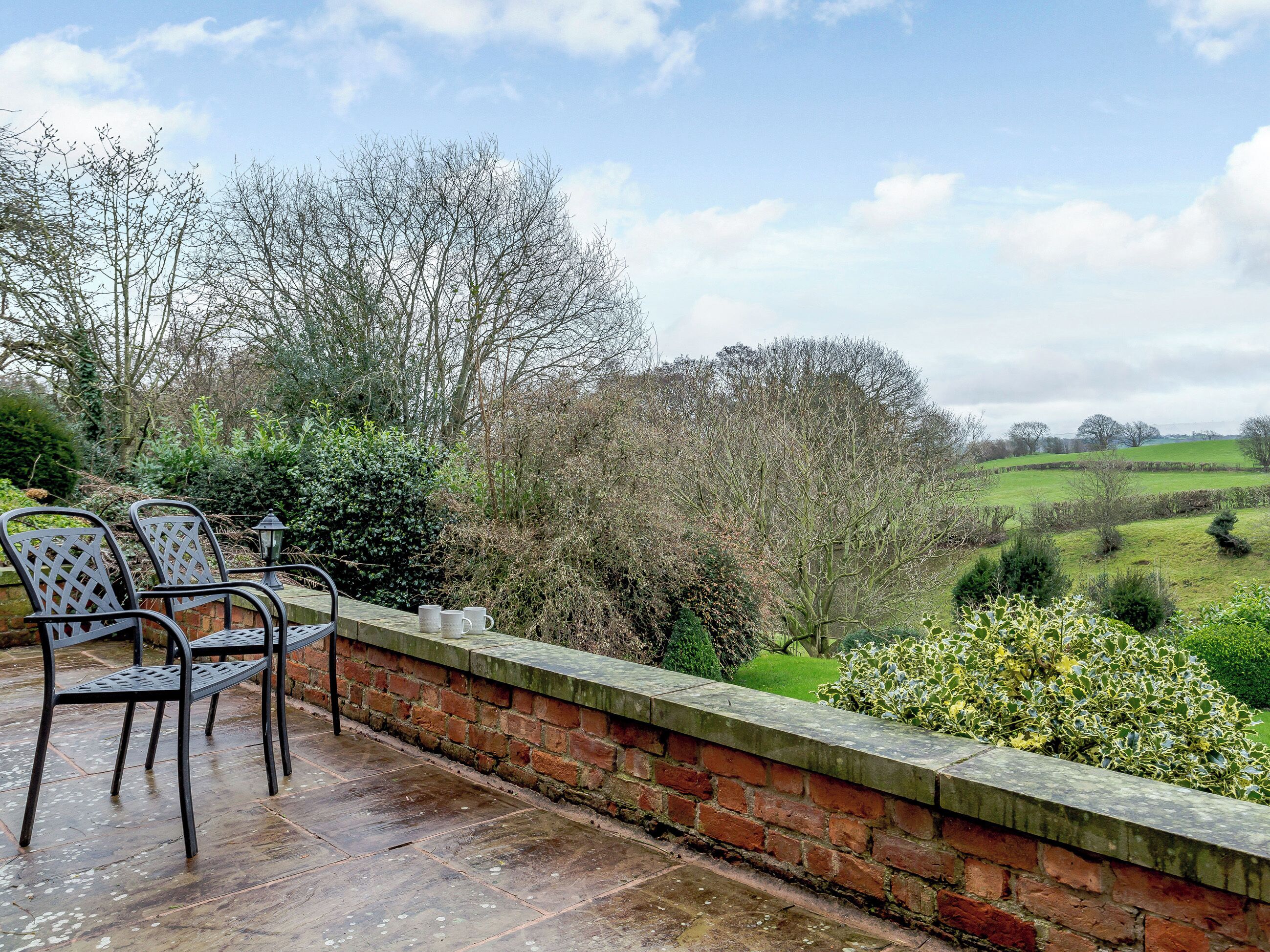 Une terrasse avec des chaises et une table, donnant sur un paysage verdoyant parsemé d’arbres et bordé d’un mur de briques.