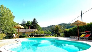 En swimmingpool med solstolar och parasoller, omgiven av grönska och med ett berg i bakgrunden.