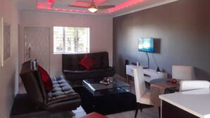A modern living room with a ceiling fan, a flat-screen TV mounted on the wall, a sofa, a coffee table, and a dining area with a table and chairs. - Marina Glen Resort No.46 (Southbroom)