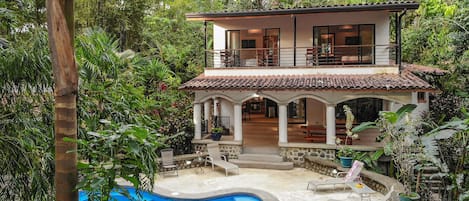Una casa de dos pisos con piscina, rodeada de abundante vegetación.