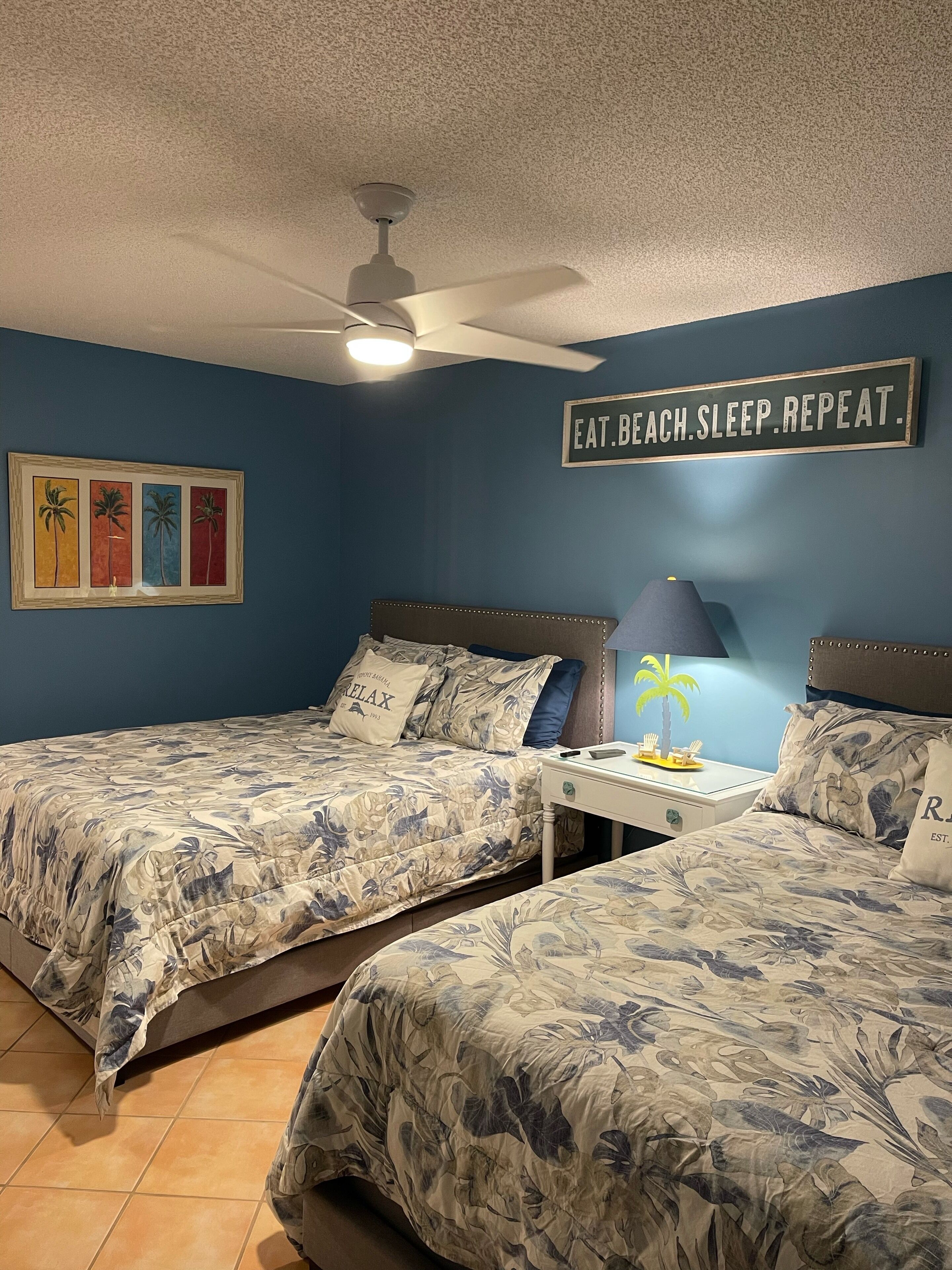 Une chambre avec deux lits, un ventilateur de plafond, une table de chevet avec une lampe et un panneau mural sur lequel on peut lire : « MANGER. PLAGE. DORMIR. RÉPÉTER. »