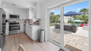 A modern kitchen with white cabinets, stainless steel appliances, and a large sliding glass door leading to an outdoor patio with a pool and umbrellas. - Tequesta Beach House, Heated Private Pool, Close to Beach, Sleeps 8 (Jupiter)