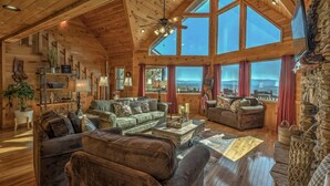 A spacious log cabin interior with multiple seating areas, large windows offering scenic views, and a stone fireplace.