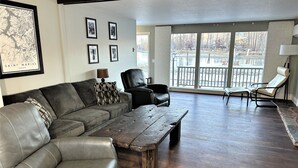 Un salon avec un canapé en cuir gris, une table basse en bois et un fauteuil en cuir noir. Des tableaux encadrés ornent le mur, et une grande fenêtre offre une vue sur un quai et des arbres.