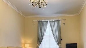 A room with a sofa, a TV on a stand, a window with curtains, and a chandelier. - A spacious house in an ideal location, across the road from the beach. (Kirkcaldy)