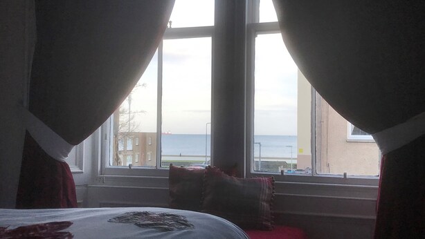 A bedroom with a large window, a bed with a white cover, and a view of the sea and buildings. - A spacious house in an ideal location, across the road from the beach. (Kirkcaldy)