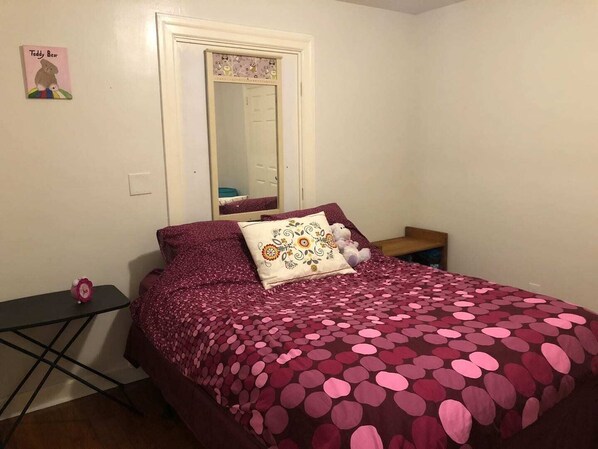 Une chambre à coucher avec un lit recouvert d’une couverture à pois, une table de chevet sur laquelle est posé un ours en peluche, et un miroir au-dessus du lit.